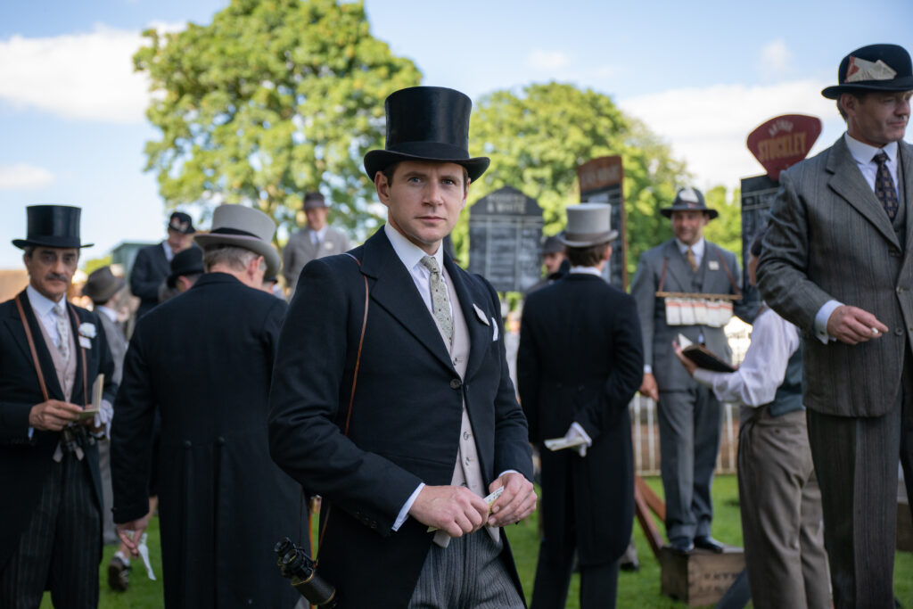 A group of men in period attire, including top hats and formal coats, gather outdoors with a focus on a well-dressed man in the foreground, amidst a lively social atmosphere.