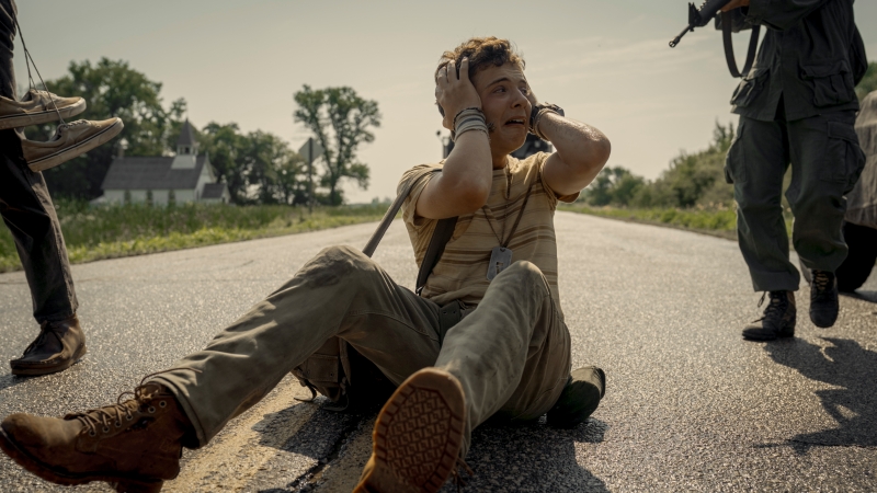 A young man sitting on a road, covering his head in distress while armed figures approach him in a tense atmosphere.