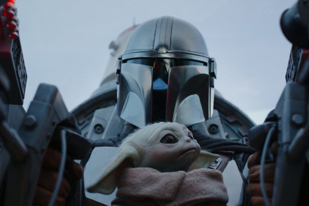 A close-up of a helmeted figure holding a small, alien creature, set against a cloudy sky.