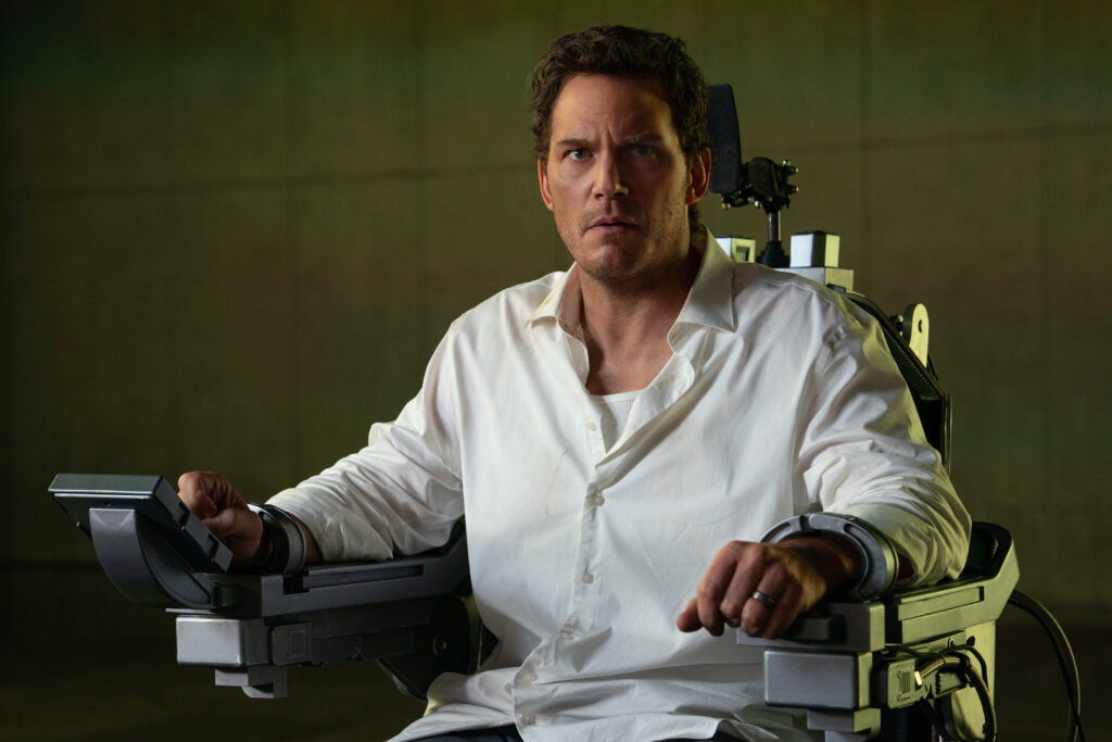 A man with a concerned expression sits in a futuristic restraint chair, wearing a white shirt, with a control panel in front of him and a green backdrop.