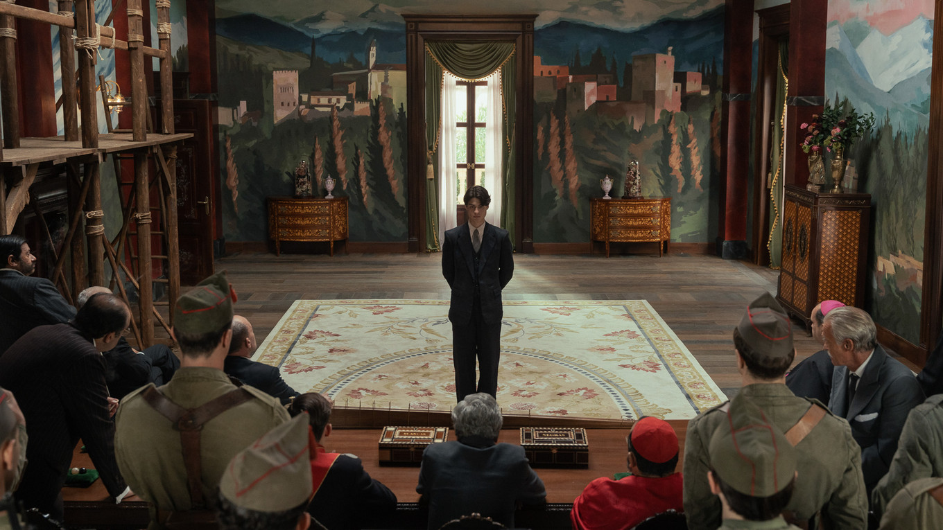 A man stands before a seated gathering in an ornate room in La Bola Negra
