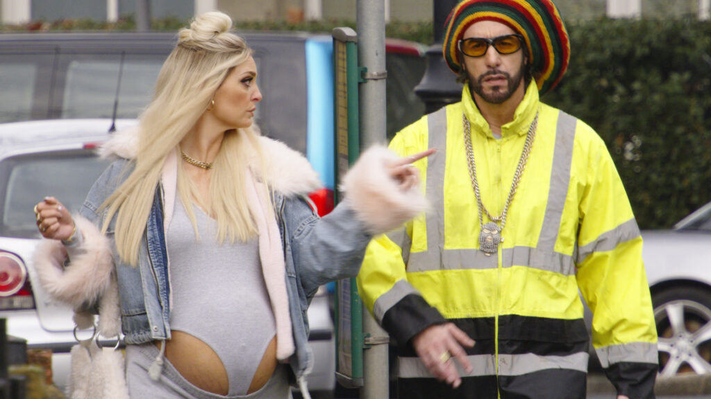 Morgana Robinson as Julie gestures towards Sacha Baron Cohen as Ali G, wearing a fluorescent jacket and knitted hat, in the official film still.