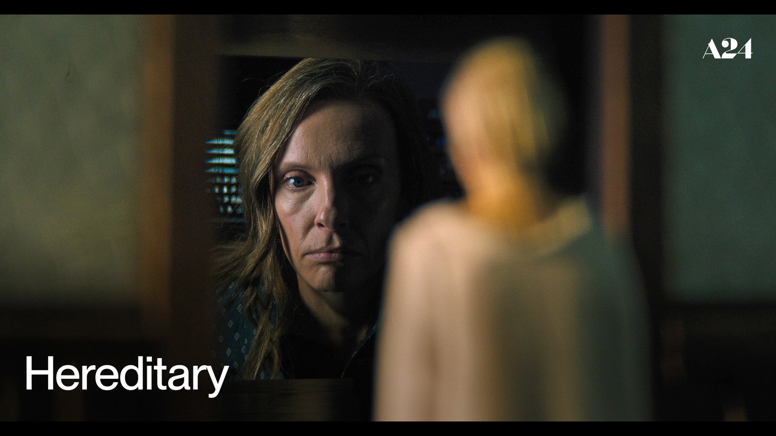 Official Hereditary artwork from A24 showing Toni Collette in a dark doorway beside a blurred figure
