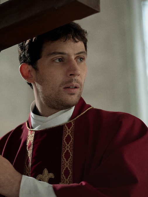 Josh O'Connor as Father Jud in red vestments holding a wooden cross in Wake Up Dead Man